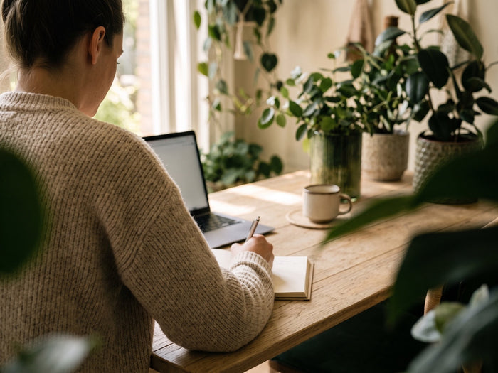Een medewerker schrijft in een notitieboek aan een houten bureau tussen de planten
